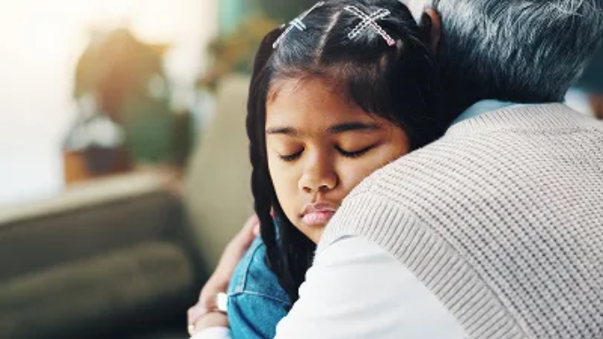 A young girl with her eyes closed leans her head against an adult's shoulder in a warm embrace.