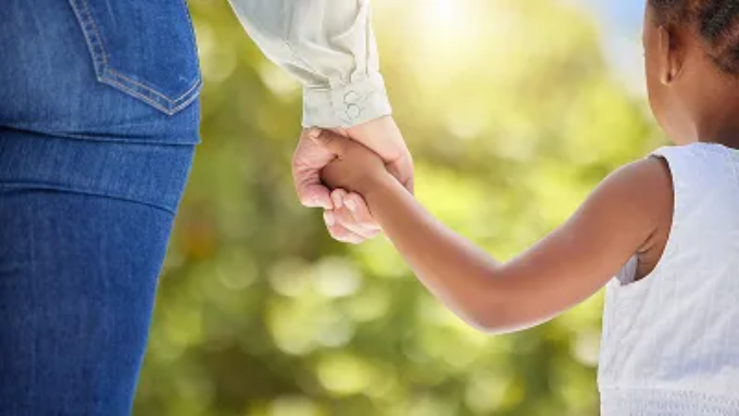 A child and an adult hold hands while walking together outdoors in the sunlight.