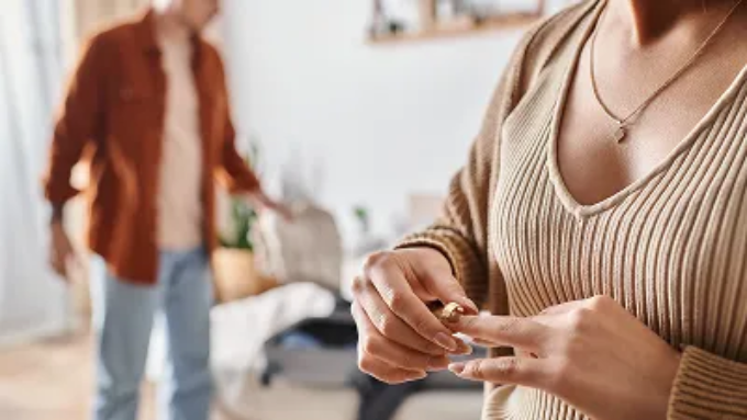 A woman removes a wedding ring from her finger while a man stands blurred in the background.