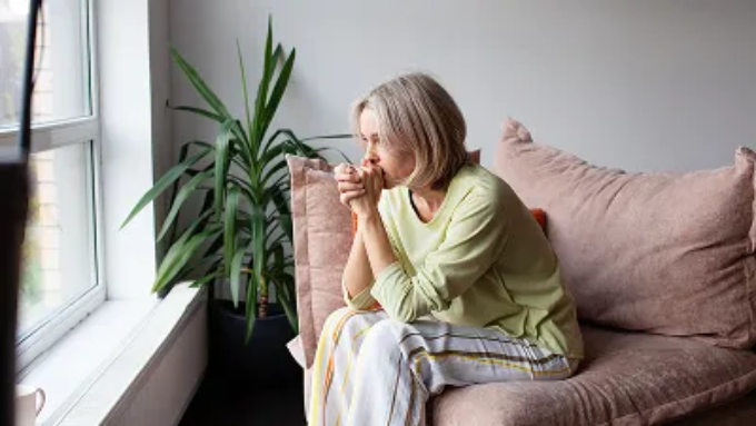 A thoughtful older woman looks out a window while sitting on a pink sofa.