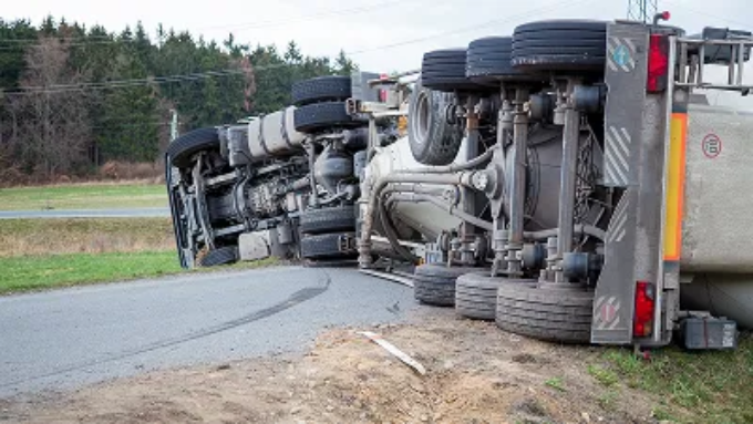 A large semi-truck and its trailer lay overturned on their side across a two-lane road.